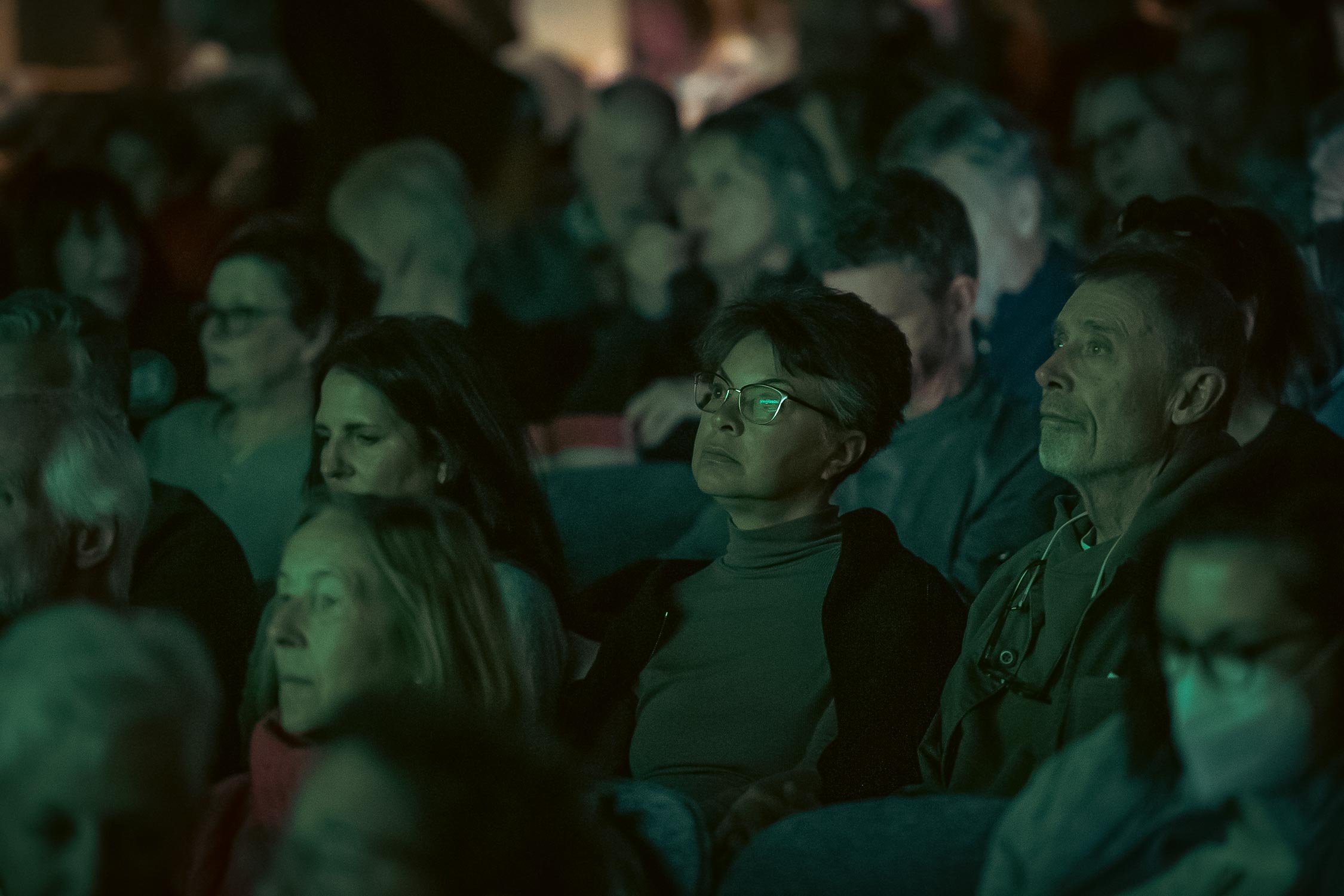 A dimly list theatre, we see the faces of a few audience members looking forward.
