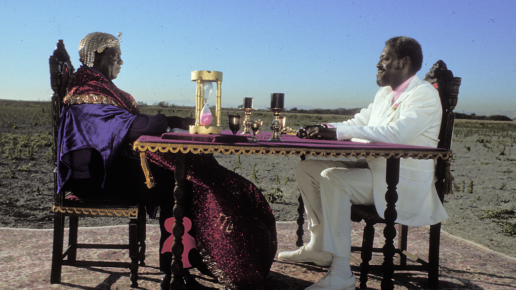 Space is the Place Still film still. Two men sit at a royal looking table facing each other. One man is in a white suit the other is in a gold head covering and royal robes. They are sitting in the middle of a desert like landscape.