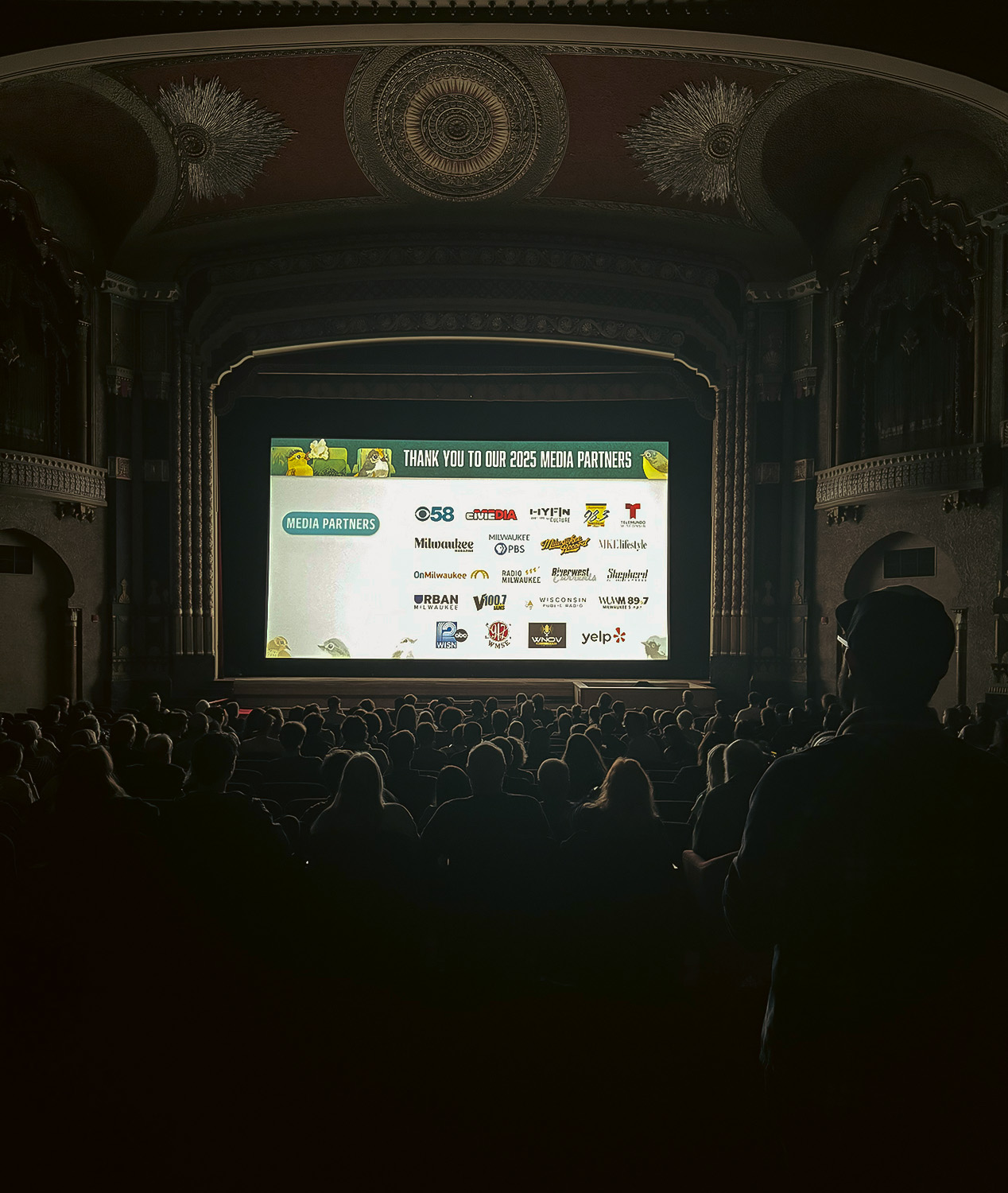 A dark theatre with a bright screen showing community partner logos.