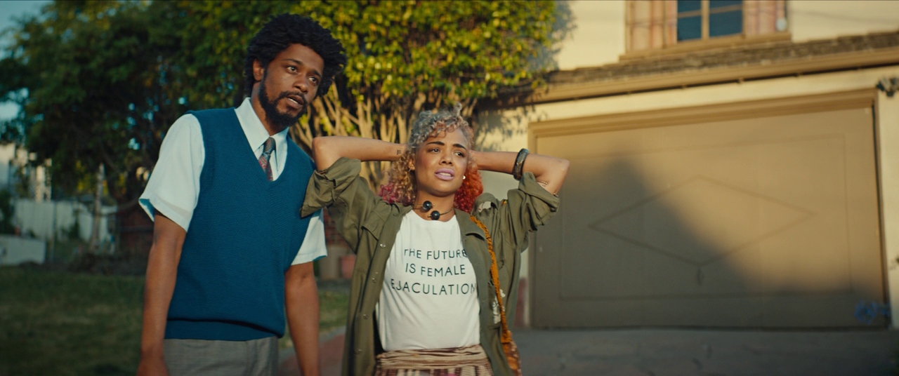 Sorry To Bother You film still. Cassius (LaKeith Stanfield) and Detroit (Tessa Thompson) stand next to each other in front of a house looking off to the side of the image.