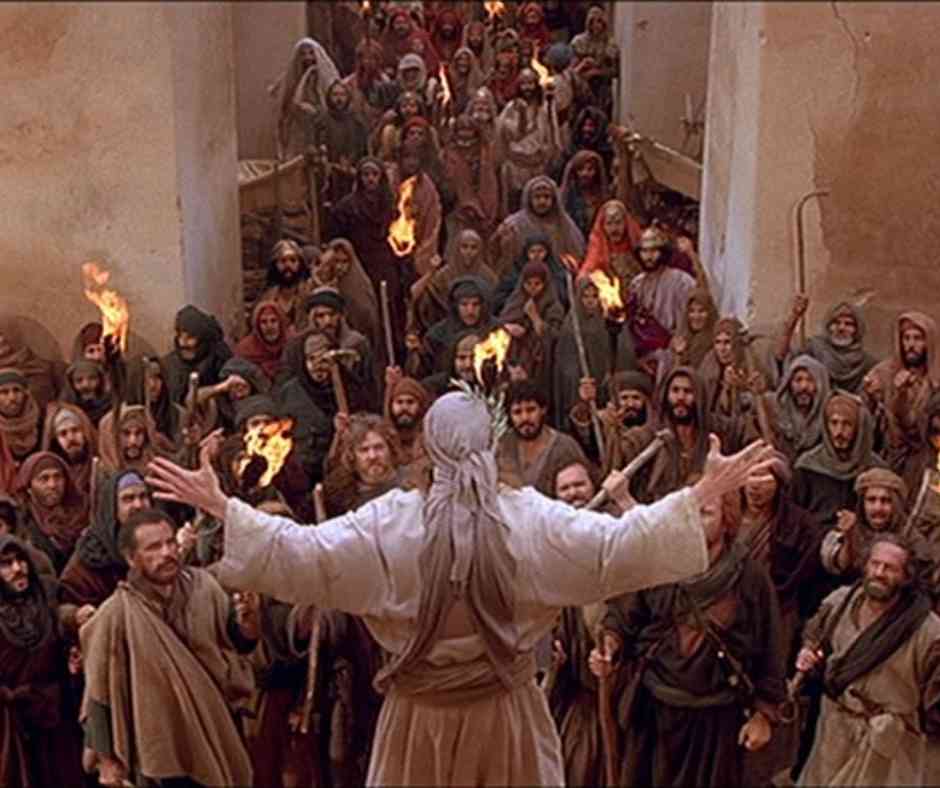 The Last Temptation of Christ Film Still The back of a man facing a large crowd of people, his arms are raised up at his sides and people in the group are holding lit torches.