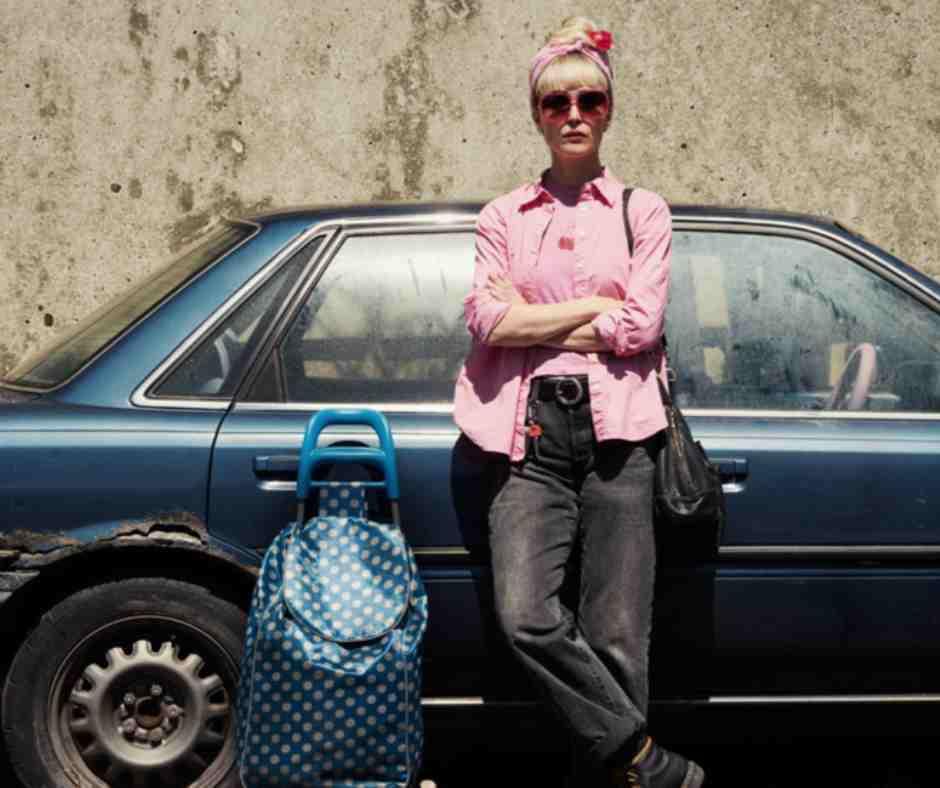 Tow Film still. A woman in a pink button up leanrs against a blue car with a big blue bag next to her on the ground.