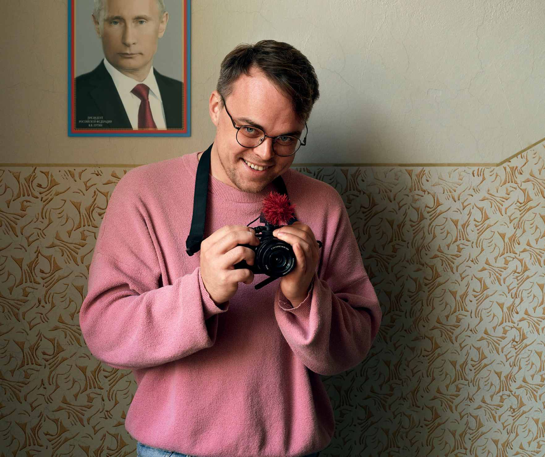 A man in a pink pullover sweater hold a camera up. A photo of Putin is hanging on the wall behind him.