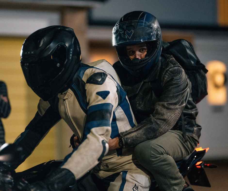 Pillion Film Still Two men on a motorcycle together, both wearing helmets. The man on the back has his helmet visor up.