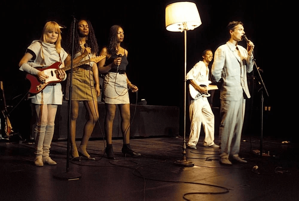 Scene from the Talking Heads film STOP MAKING SENSE. The image features singer David Byrne singing while holding a lamp, and the background singers and bass player standing to his right.
