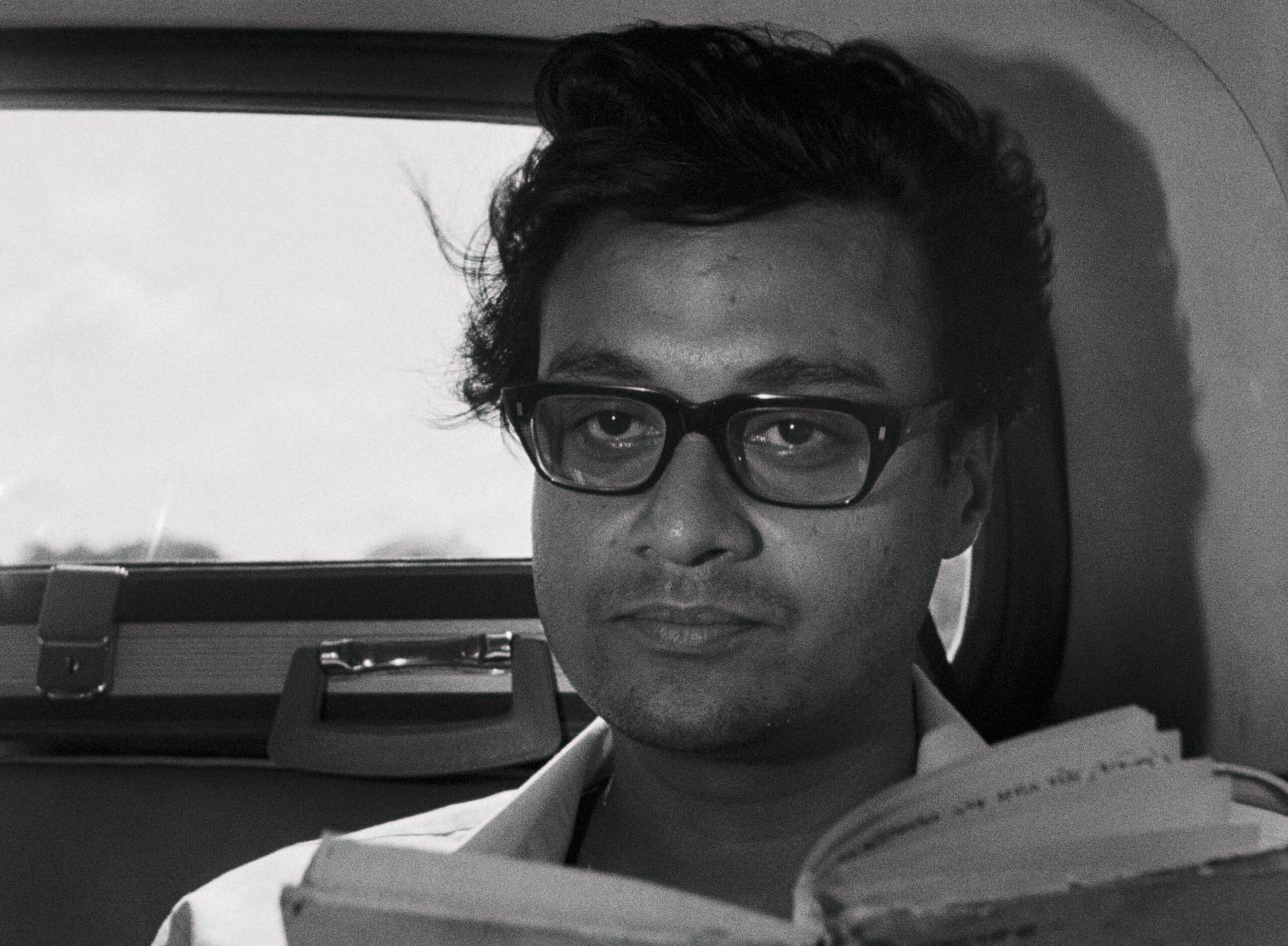 Days and Nights in the Forest film still. A black and white portrait of a man with glasses, holding a book while sitting in a car.