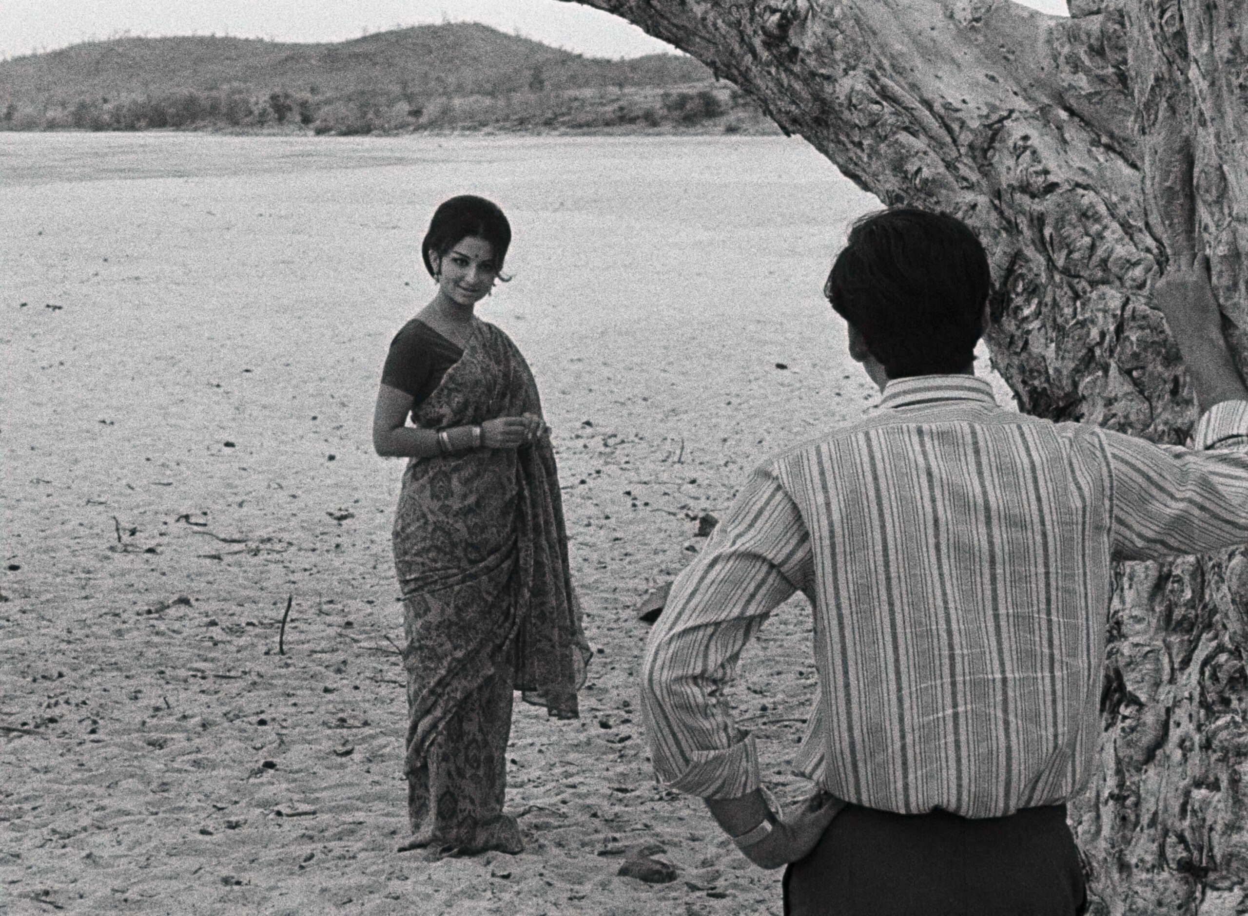 Days And Nights In The Forest Film Still A woman with her hair in a neat updo faces a man with his back towards the camera and his hand on a tree that stands between them.