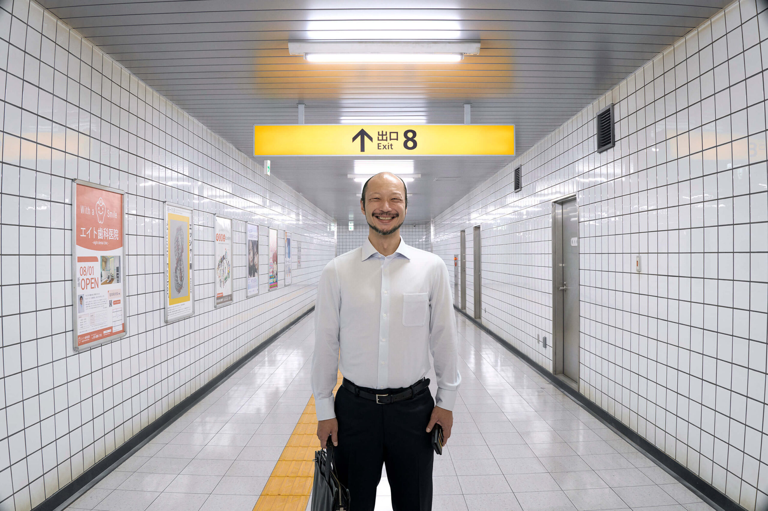 Exit 8 Film Still A man stands facing forward, smiling.