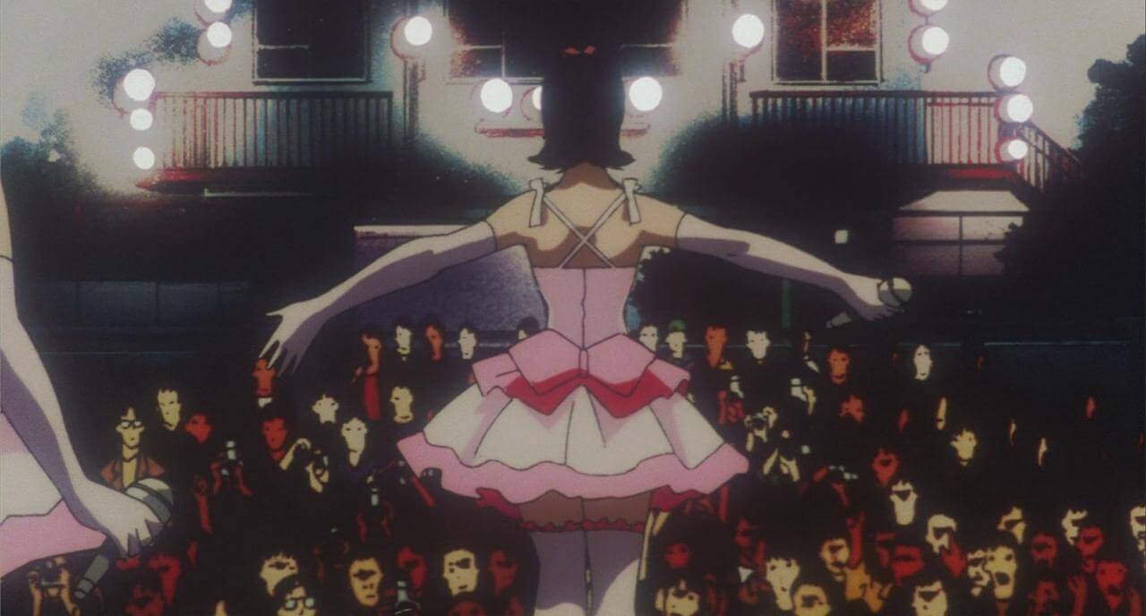 Perfect Blue Film Still A woman in a pink dress faces towards a full audience looking up at her.