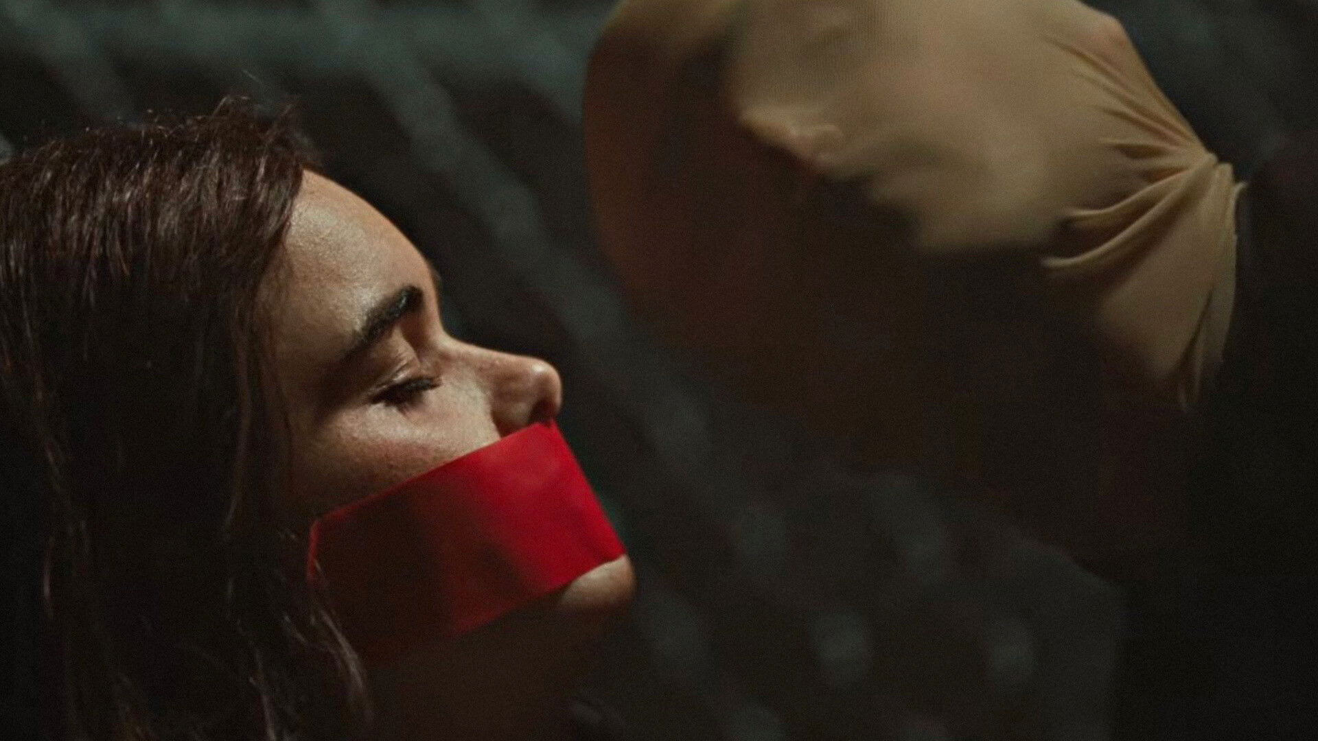 Faces of Death Film Still A woman has her eyes closed with a red piece of fabric covering her mouth. A masked face stairs back at her.