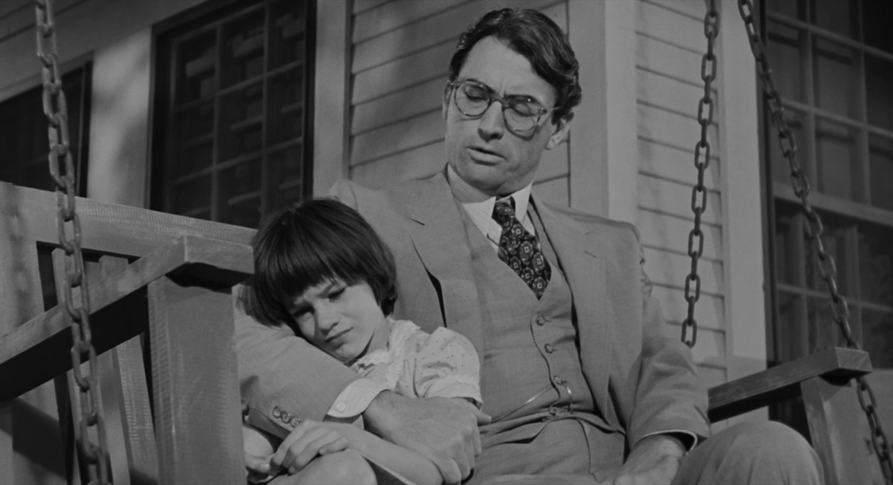 To Kill A Mockingbird A father and his child sit in an embrace on a porch swing, the child's face looks troubled and the father looks to be comforting them.