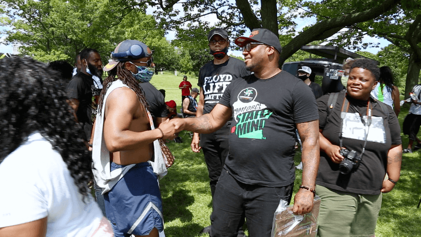 A still from the film Heavy is the Crown, featuring a crowd of people engaging with a community activist in a park.