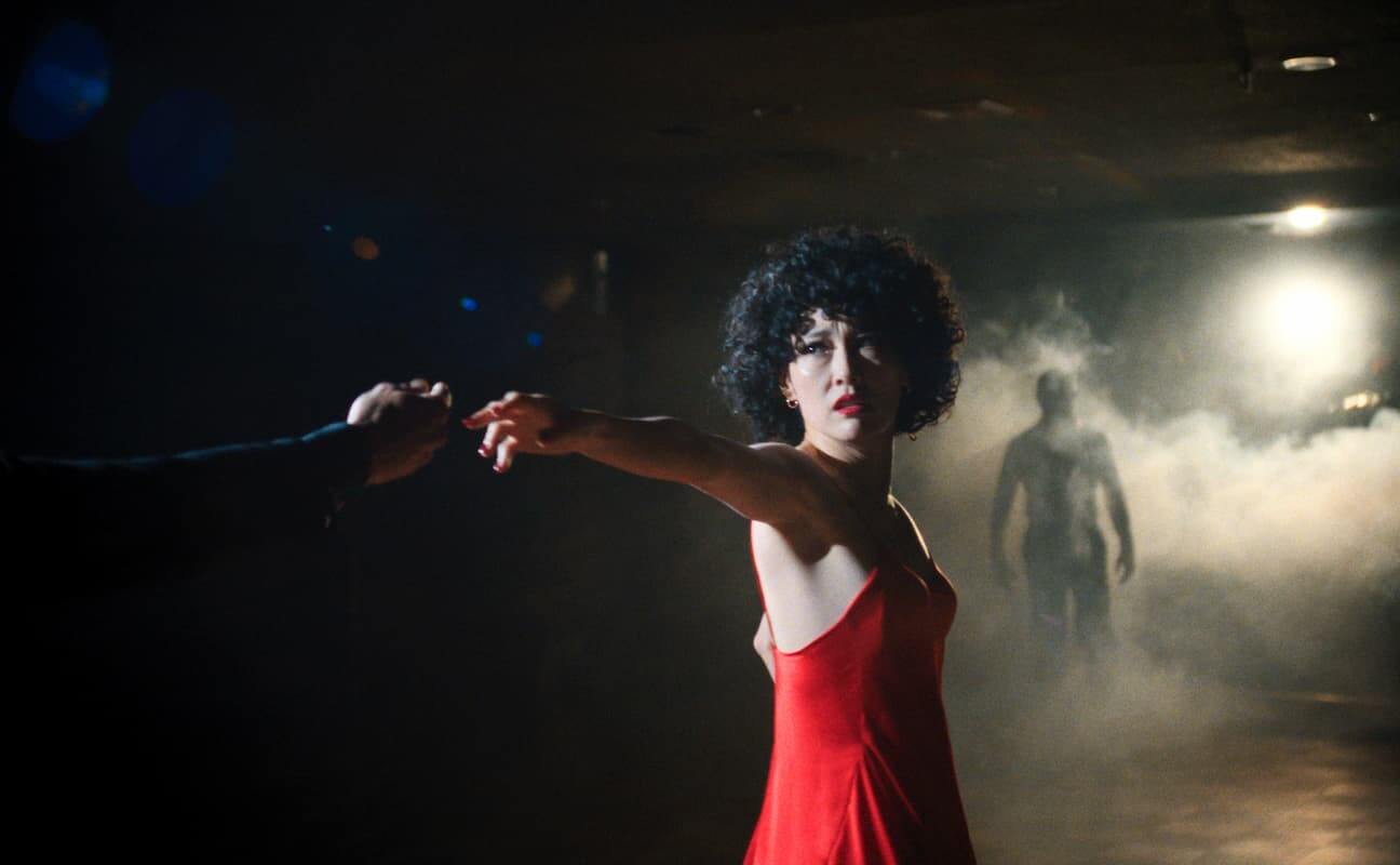 Ha-Chan Shake Your Booty Film Still A woman with short tight curly hair in a red dress extends her arm holding a pistol.