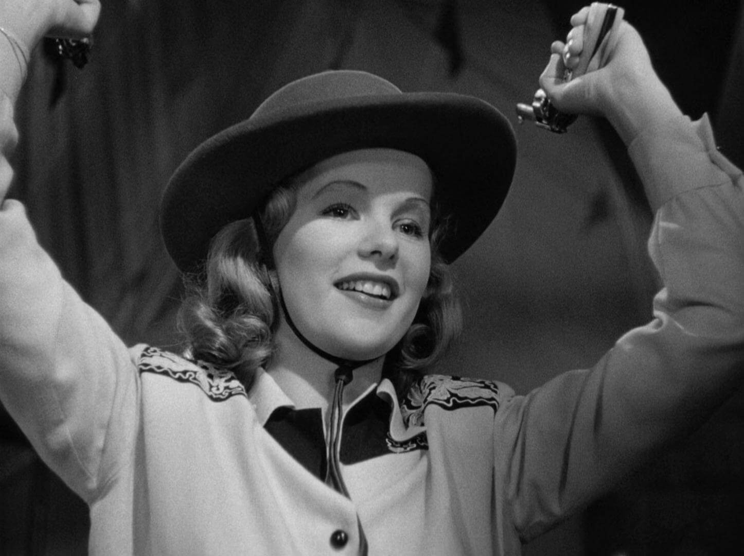 Gun Crazy Film Still Black and White a woman in a uniform and brimmed hat stands with her arms above her head smiling